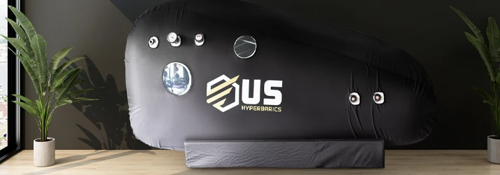 Large black chamber with circular ports and a “US Hyperbarics” logo, positioned between two potted plants.